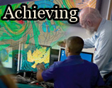 Two meteorologists analyze weather data on computer screens with a large weather map behind them. The image contains the text "Achieving".