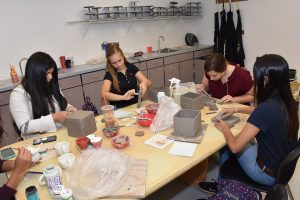 Students collaborate on pottery projects in a bright art classroom. Four young women work with clay, shaping pots and boxes. Supplies surround them on the table, with sinks and storage in the background.