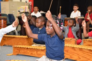 A young Black girl plays a xylophone with enthusiasm in a music class, surrounded by other children holding mallets. The classroom features wooden xylophones and a file cabinet.
