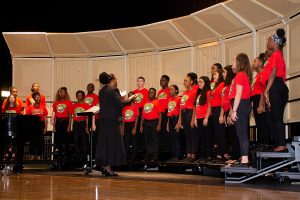 A diverse youth choir, wearing matching red t-shirts, performs on a stage with a conductor leading them. A pianist is visible to the left.
