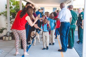 Elementary school students, including a young boy in a blue shirt, receive high fives from adults and a therapy dog as they enter the school. A line of uniformed officers stand in the background.