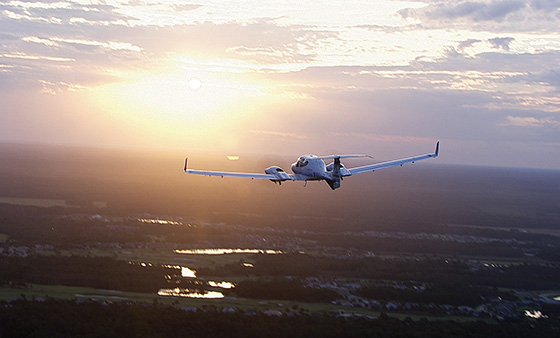 Diamond DA42 Twin Star soars above a lush landscape at sunset. The sun illuminates the aircraft, casting a warm glow over the trees and water below.