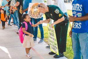 A young girl in a school uniform fist-bumps a police officer holding a sign that reads "Be a Treasure Hunter, Search for what's talents, skills and intelligence lie." Other adults hold signs nearby.