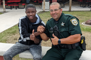 A smiling Black teenager and a uniformed sheriff's deputy give thumbs up while sitting together outside. The deputy wears a green uniform with a badge. They clasp hands in a gesture of camaraderie.