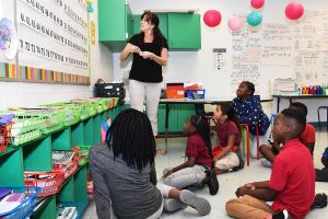A teacher instructs a diverse group of elementary students on the floor in a colorful classroom. Math concepts are visible on the whiteboard, and learning materials are organized in colorful bins.