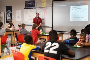 A diverse group of middle school students sit at desks in a classroom, listening to a male teacher present a "Decision Matrix" on a projector screen. The teacher wears a red polo shirt.