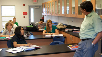 A high school science class in session: students at desks listen to their teacher lecturing in a bright classroom, with lab counters and cabinets visible.