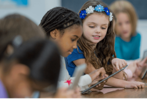 Elementary school students, including a girl with a floral headband, work together on tablets in a classroom setting, fostering collaborative learning.
