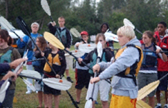 Group of kids outdoors holding kayak paddles, practicing strokes on land before getting in the water, wearing life vests, learning kayaking techniques.