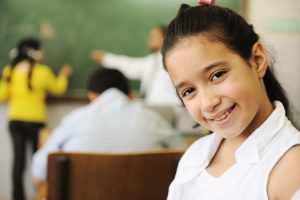 Smiling young student with dark hair and a pink bow, in a white top, looks directly at the camera in a classroom setting. Other students and a chalkboard are blurred in the background.