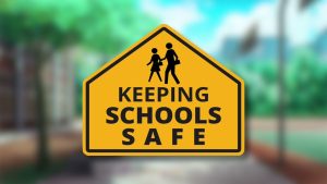 Yellow road sign with a black border reads "Keeping Schools Safe" above a silhouette of an adult and child walking. A blurred school building and trees are in the background.