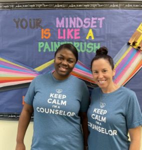 Two smiling school counselors stand in front of a bulletin board that reads "Your Mindset Is Like A Paintbrush." They wear matching blue t-shirts that say "Keep Calm I'm A Counselor.