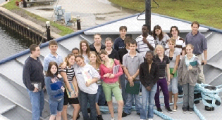A diverse group of high school students and two adults pose on the deck of a canal boat. They are smiling and holding notebooks, with a canal lock visible behind them.