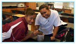 Two smiling Black high school students in a science classroom examine a specimen in a container, likely for a biology or environmental science project.