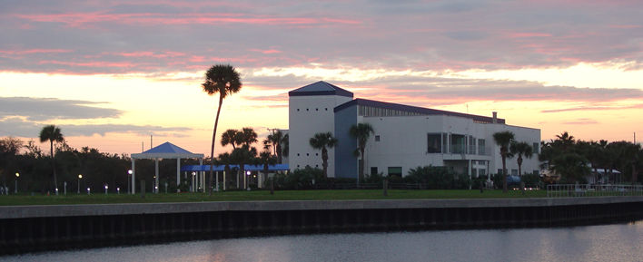 Waterfront building at sunset with palm trees, blue accents, and a covered walkway. Pink and orange hues fill the sky, reflecting on the water.