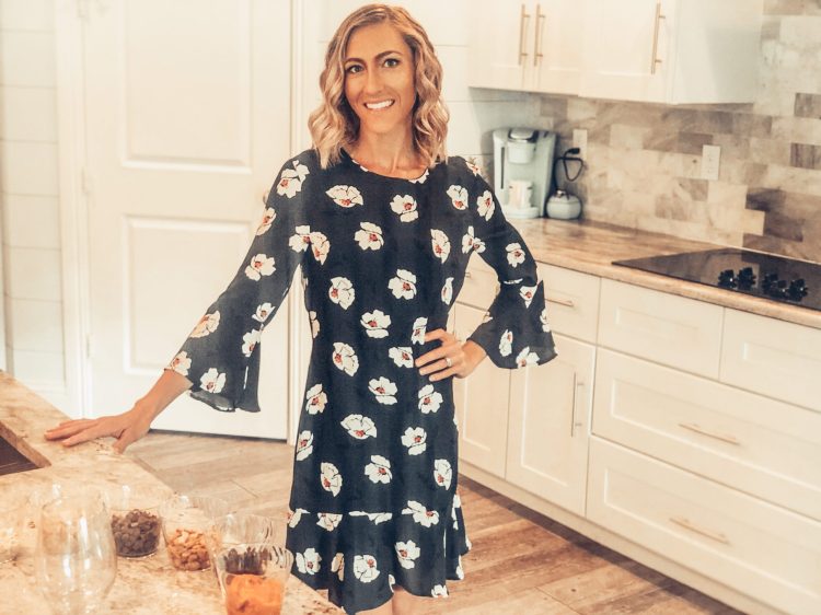 Woman in a floral dress smiles in a bright kitchen, leaning on a counter with jars of snack ingredients. White cabinets and modern appliances fill the background.