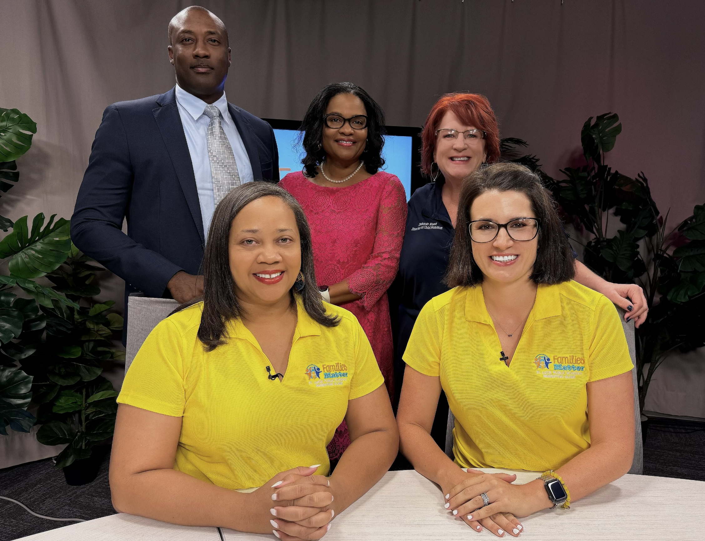 Group of five adults smiling in a studio with tropical plants